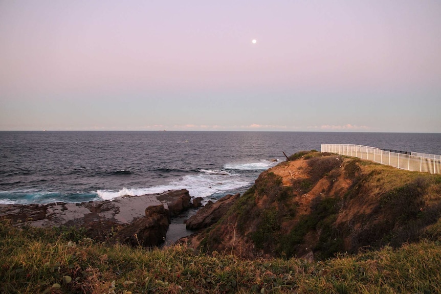 Modern photo at sunset as the moon rises about the ocean horizon about the ocean pool below. A white fence sits at the cliff top