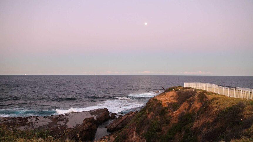 Modern photo at sunset as the moon rises about the ocean horizon about the ocean pool below. A white fence sits at the cliff top