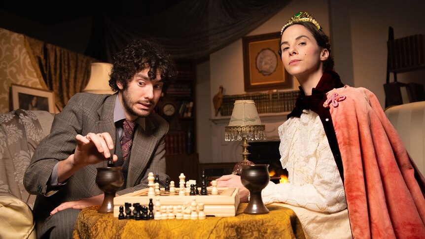 Justina dressed as a monarch and wearing a crown plays chess against Alex who is attempting a sneaky move.