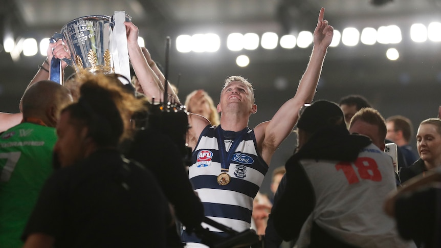 Geelong captain Joel Selwood smiles and looks up into the lights he wears a premiership medal and holds the cup in one hand. 