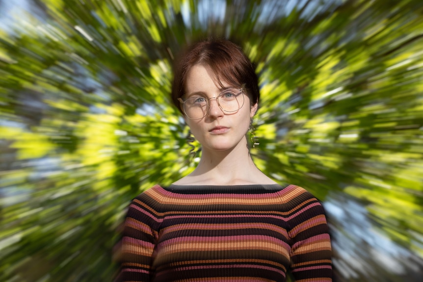 A woman stares at the camera, the background of trees blurry and whirling behind her