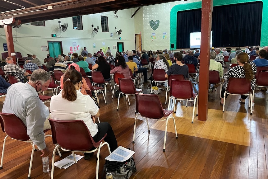 A lot of people sitting on chairs, wearing masks inside a building.