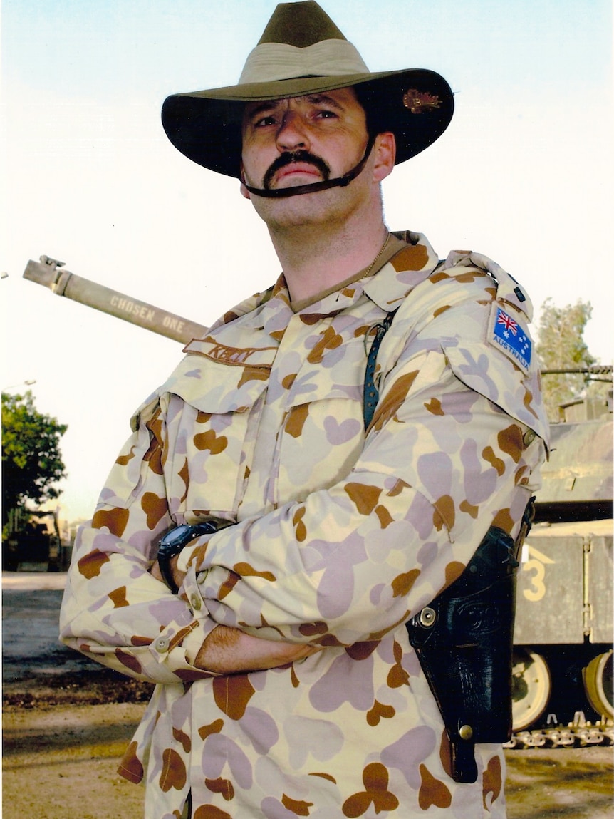 A moustachioed man in military fatigues and a hat with one of those weird chin straps stands with his arms crossed.