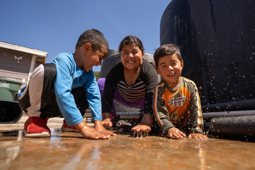 Three small children with their hands in water spilled on the ground grin at the camera