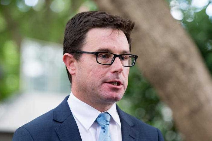 A bespectacled man wearing a navy suit, white shirt and pale blue tie speaking with a tree trunk and leaves in the background