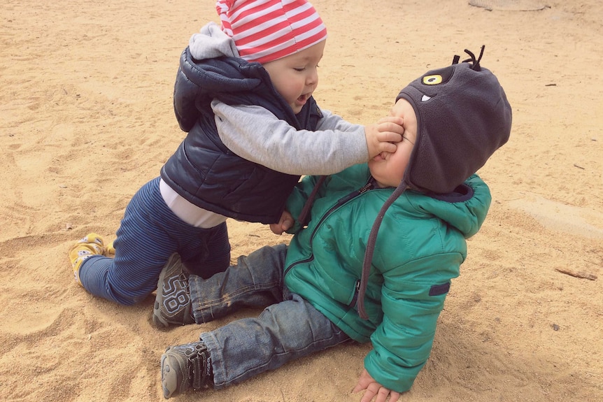 Two toddlers dressed in warm clothes in a sandpit. One leans over the other, roughly pushing their hand into the other's face.