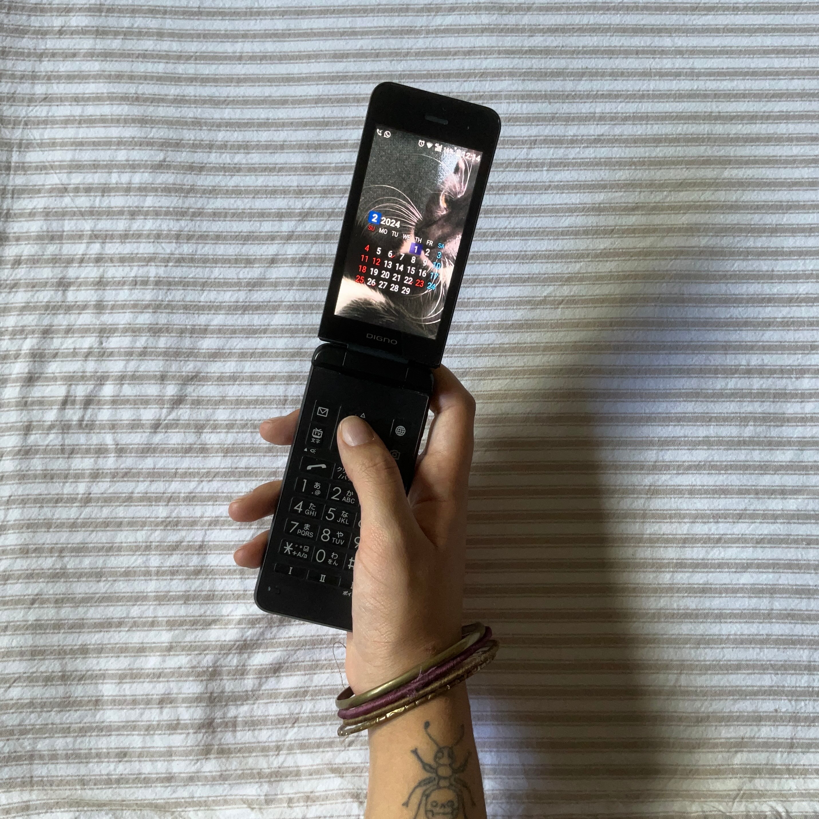 A close up of a hand holding and using a flip phone above the surface of striped bed sheets.