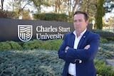 Paul Toole stands with his arms crossed in front of Charles Sturt University entrance sign in Bathurst.