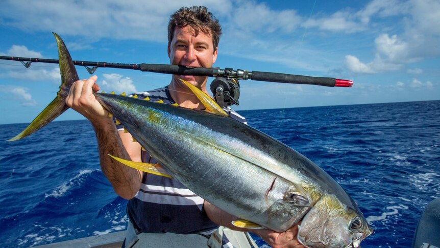 Clarke Gayford holds a massive fish in both hands, while balancing a fishing rod in his mouth