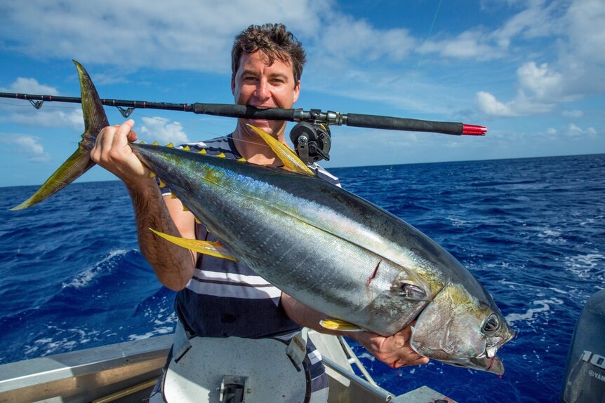 Clarke Gayford holds a massive fish in both hands, while balancing a fishing rod in his mouth