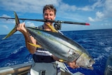 Clarke Gayford holds a massive fish in both hands, while balancing a fishing rod in his mouth