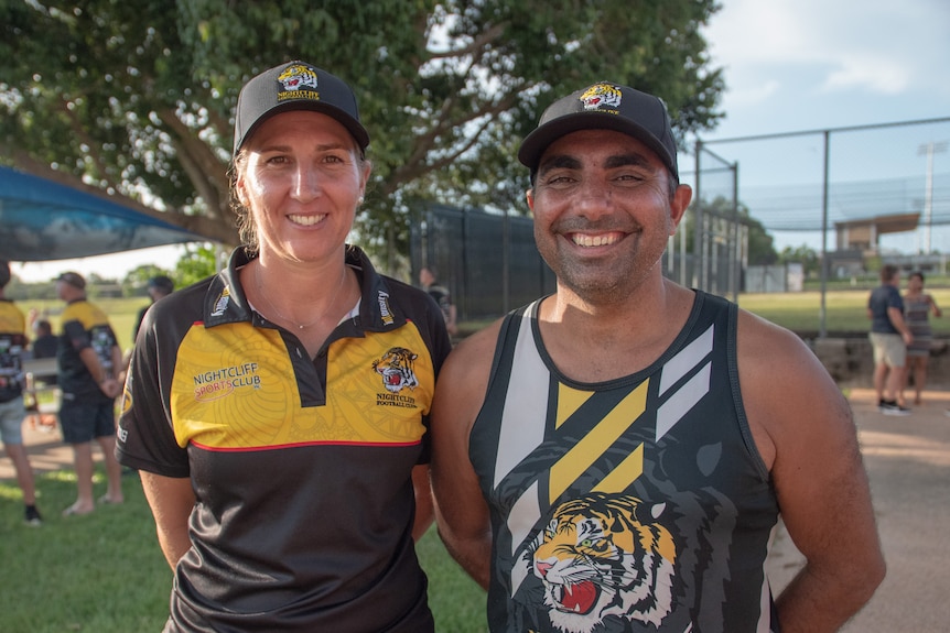A woman and a man stand next to each other wearing footy club shirts and smile at the camera
