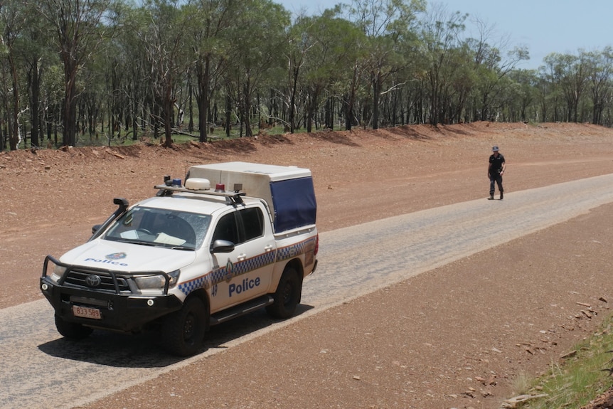 Police officer walks along a stretch of a remote highway with a police car in the foreground