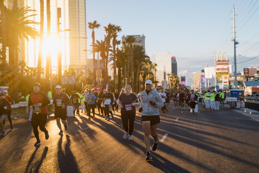 A crowd of runners run on the street, palm trees, a big M and sun rises behind them. 