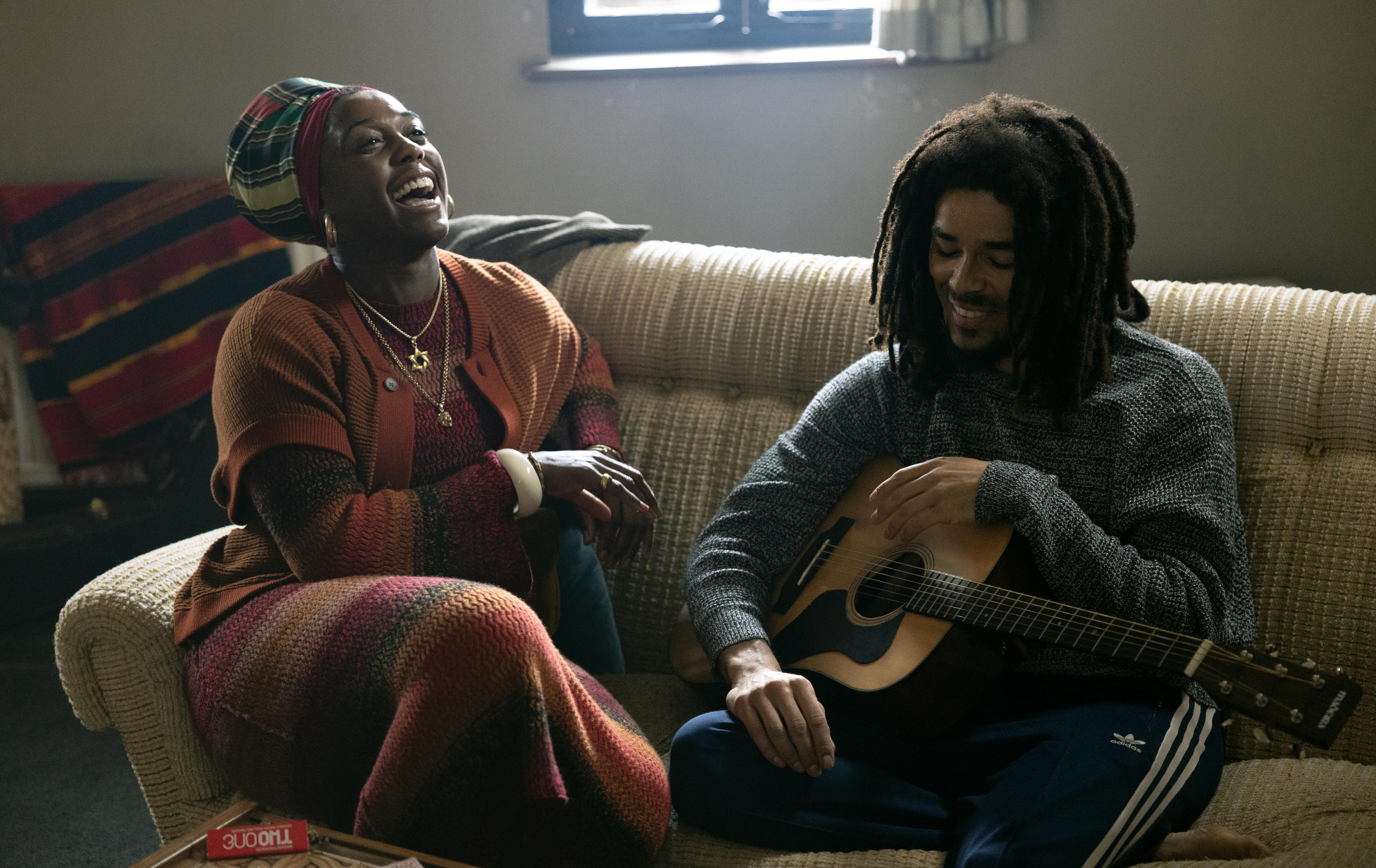 Two actors, one holding a guitar, the other laughing, sitting together on a couch