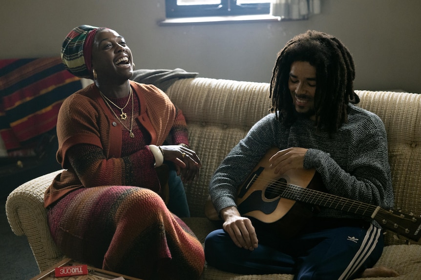 Two actors, one holding a guitar, the other laughing, sitting together on a couch