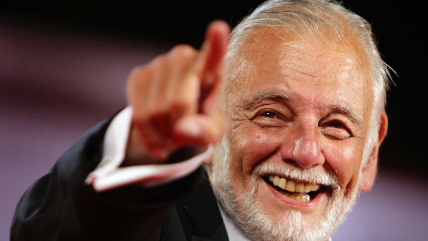 George A Romero, sporting a short grey beard and slicked sliver hair, smiles and points towards photographers.