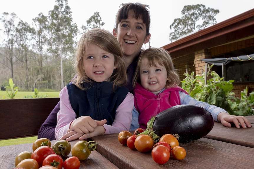 A woman holding two young girls in her lap sits at a backyard table, with eggplant and tomatoes.