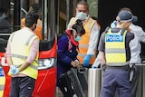 A woman wearing a surgical mask carrying a suitcase gets off a red bus while surrounded by security and police.