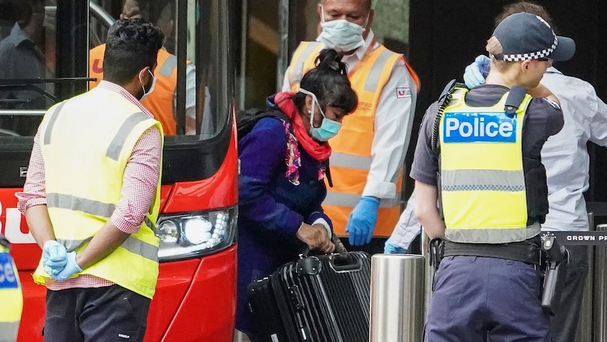 A woman wearing a surgical mask carrying a suitcase gets off a red bus while surrounded by security and police.