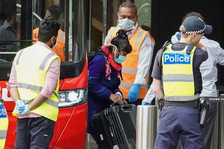 A woman wearing a surgical mask carrying a suitcase gets off a red bus while surrounded by security and police.