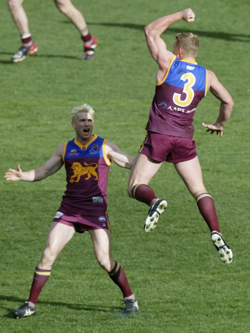 A Brisbane Lions AFL player leaps high in the air in celebration as his teammate looks back at him during a grand final.