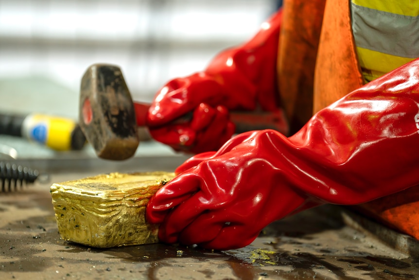 A bright gold bar, with two hands in long red gloves, one holding a hammer