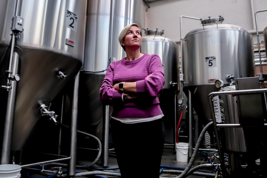Woman with blonder hair wearing purple cardigan stands in brewery with large steel containers behind