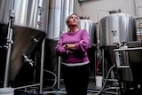 Woman with blonder hair wearing purple cardigan stands in brewery with large steel containers behind
