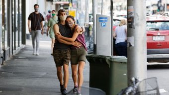 A couple wearing masks, shorts and singlets hug as they walk along a Melbourne city street.