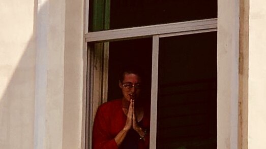 A woman wearing a red shirt holds her hands together while standing at a window