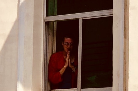 A woman wearing a red shirt holds her hands together while standing at a window