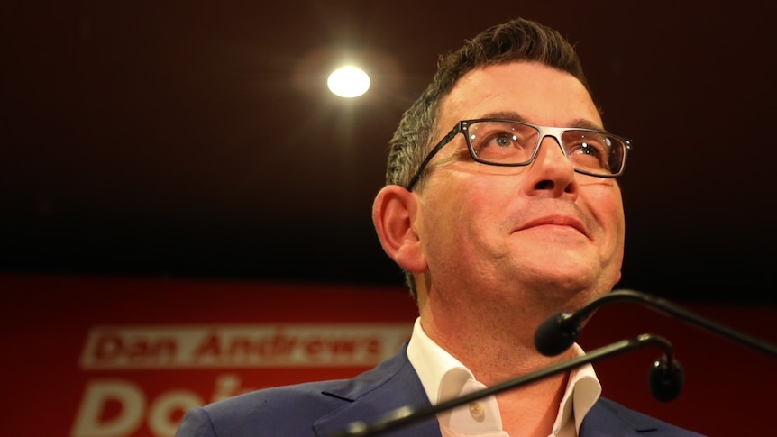 Daniel Andrews smiles, standing uplit at a podium.