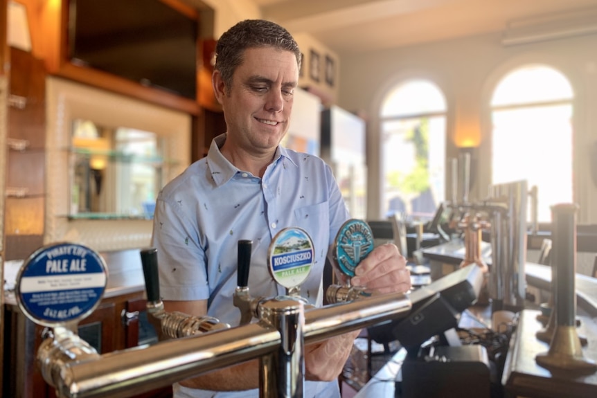 A man pulls a beer from a tap in a bar with sunlight streaming through large windows.