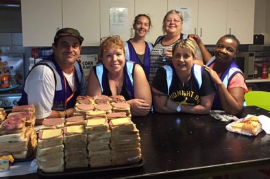 A group of people leaning on a kitchen bench with piles of sandwiches in the foreground.