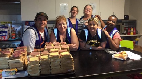 A group of people leaning on a kitchen bench with piles of sandwiches in the foreground.