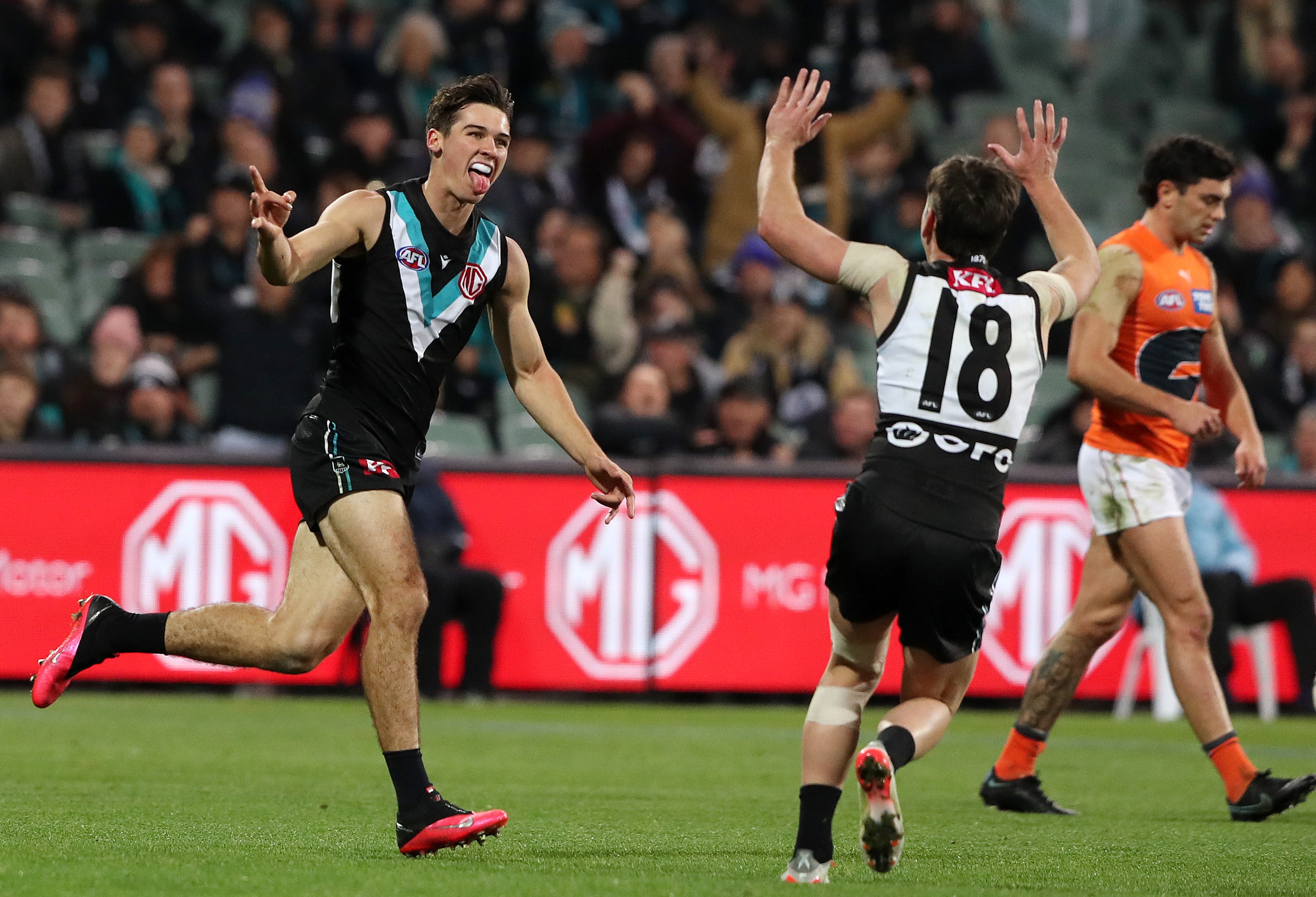 A Port Adelaide AFL player sticks his tongue out and points as he runs toward a teammate in celebration after a goal.