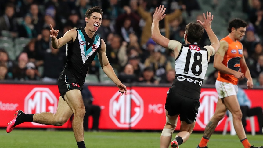 A Port Adelaide AFL player sticks his tongue out and points as he runs toward a teammate in celebration after a goal.