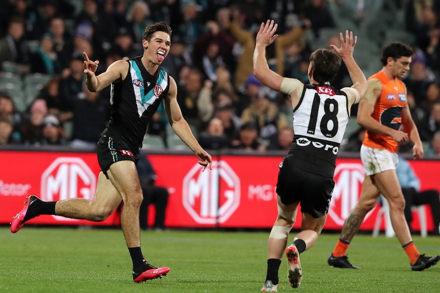 A Port Adelaide AFL player sticks his tongue out and points as he runs toward a teammate in celebration after a goal.