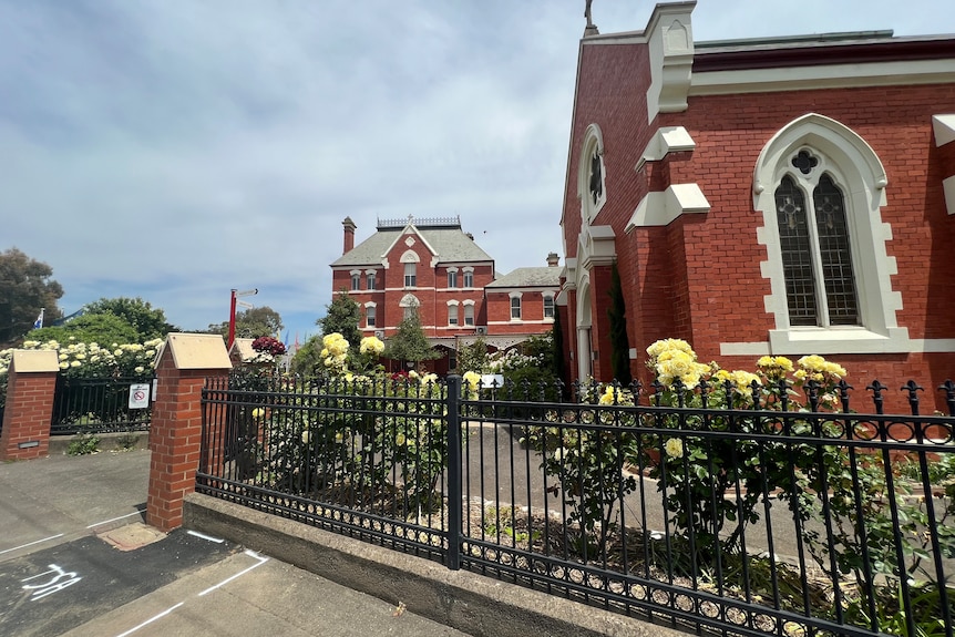 School sign that says Sacred Heart College with old red buildings behind.