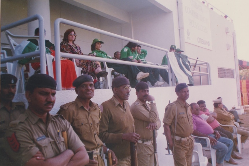 Pakistan players wear their green uniforms and sit on chairs on a balcony, as six guards stand below