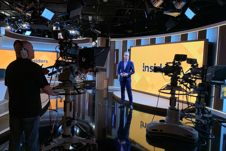 Speers in TV studio surrounded by two cameras standing in front of screen with yellow Insiders logo.