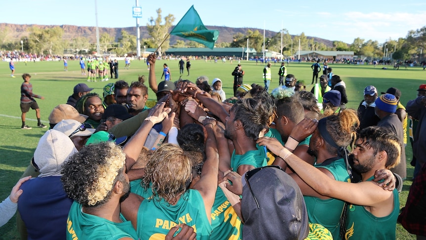Players wearing green and gold jersey stand in a group huddle after winning. 
