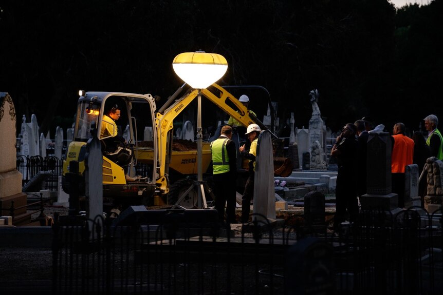 An excavator and several workers stand around a grave lit with a bright light