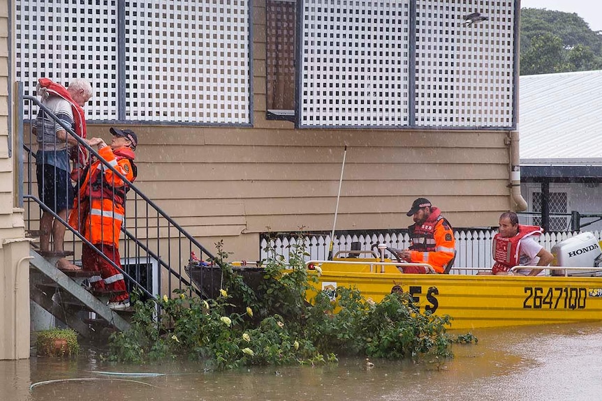 A boat waits outside a house as one person in emergency gear helps a man fit his life jacket. Water is up to the house windows