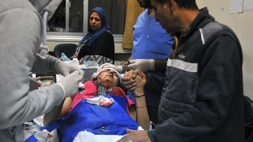 A woman is is laying down and has bleeding around her head; several people are surrounding her, including a medical worker