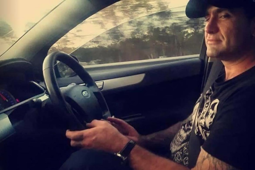 A man in a dark cap and T-shirt sits behind the wheel of a car.