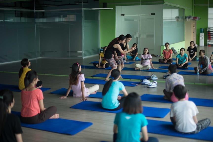 A group of women sit on gym mats while an instructor demonstrates self defence