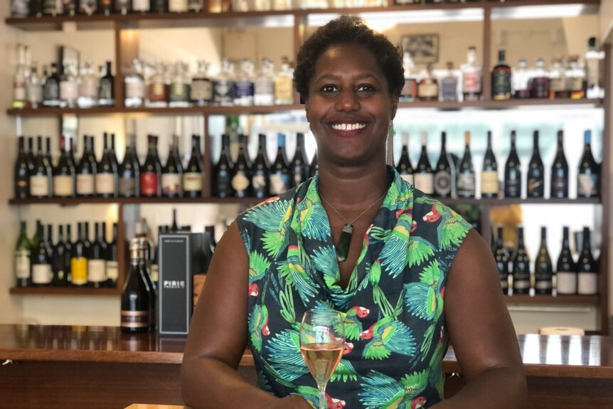 A woman standing in front of a bar holding a glass of wine smiles at the camera