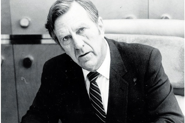 Former WA premier Ray O'Connor sits at a desk writing on a piece of paper wearing a suit.